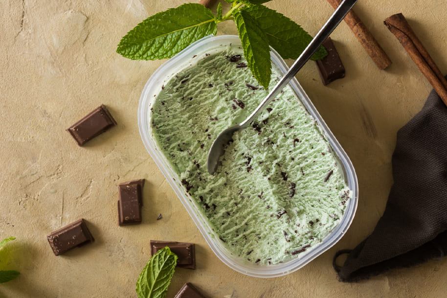 Homemade mint chocolate chip ice cream in a container, garnished with fresh mint leaves and surrounded by chocolate pieces and cinnamon sticks. The creamy texture and vibrant mint color make it a refreshing vegan comfort food option.