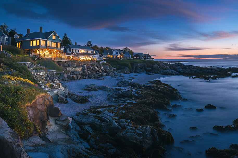 Luxury seaside houses lit up during blue hour in Ogunquit, Maine, with a rocky shoreline and calm ocean.