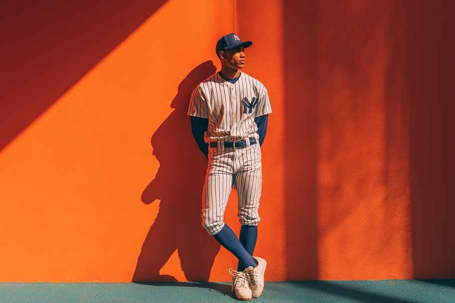 Young baseball player in striped uniform leaning casually against a bright orange wall, bat-ready pose in daylight.