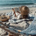 Woman reading a book on a beach blanket with a straw hat, next to a hammock and ocean waves.