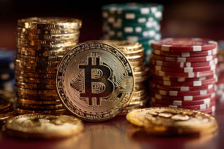 Gold Bitcoin standing upright in front of stacked poker chips and coins on a casino table.