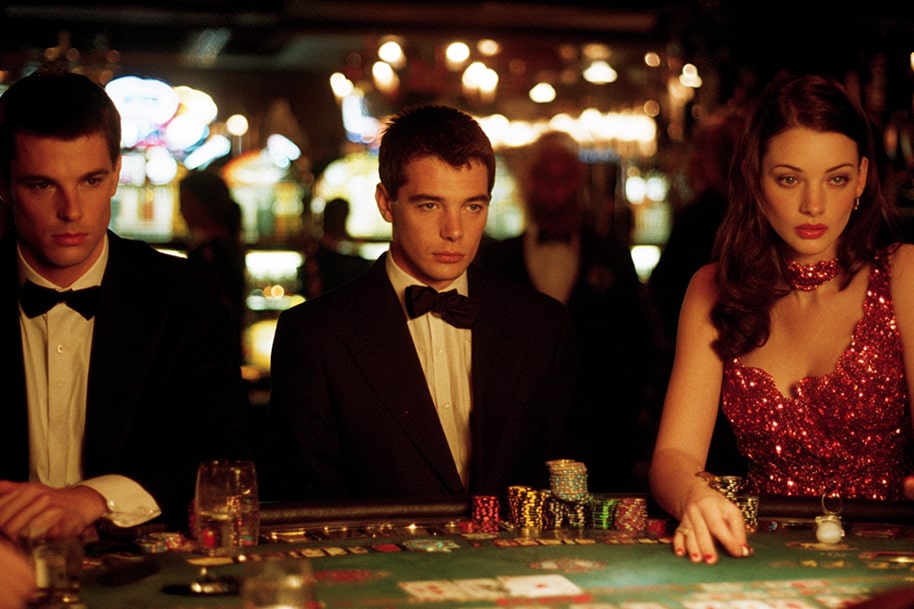 An elegant scene from the casino-themed movie “21” with three players at a blackjack table. The focus is on the casino glamour and serious facial expressions, with the sparkling slot machines in the background.