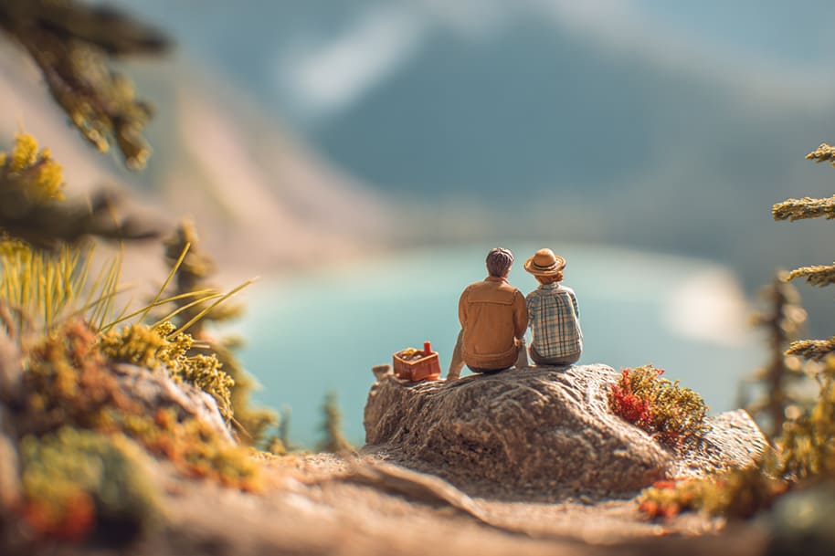 Two people sit on a rocky cliff with a picnic basket, enjoying a panoramic view of a turquoise lake surrounded by mountains, perfect for last-minute weekend getaways.