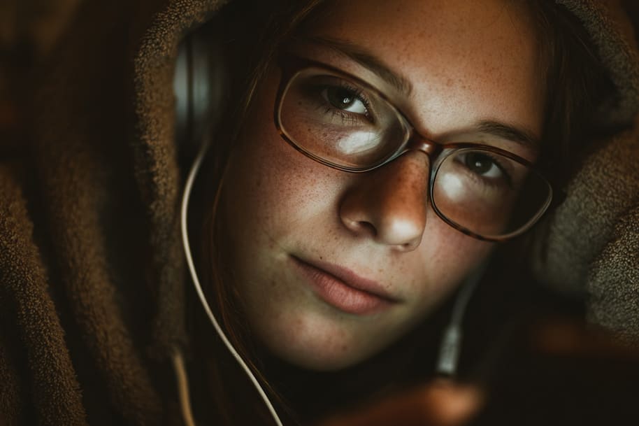 Close-up of a teenage girl in glasses and headphones, wrapped in a hoodie, looking at her phone illustrating social media’s impact on mental health and emotional well-being.