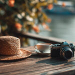Straw hat, coffee, and vintage camera on a wooden table by the water — perfect setup for bleisure travel tips and combining work with vacation relaxation.