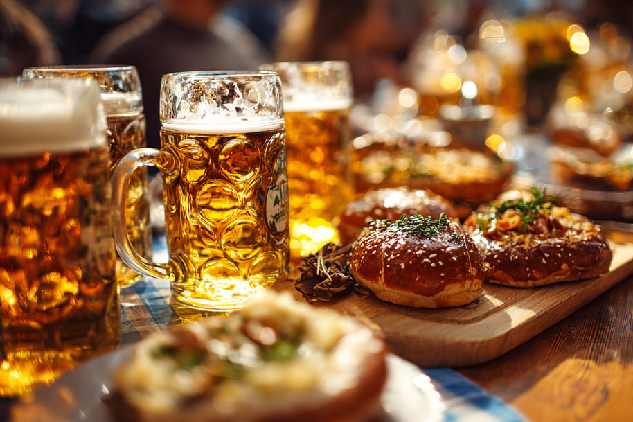 German Oktoberfest table with fresh pretzels, Bavarian pastries, and large mugs of golden beer under the sun.