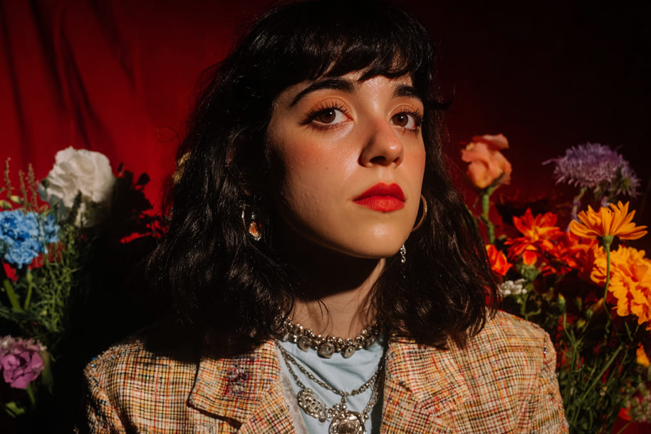 Woman with dark wavy hair and red lipstick posing against a red backdrop with colorful flowers, wearing layered jewelry and a patterned jacket.