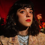 Woman with dark wavy hair and red lipstick posing against a red backdrop with colorful flowers, wearing layered jewelry and a patterned jacket.