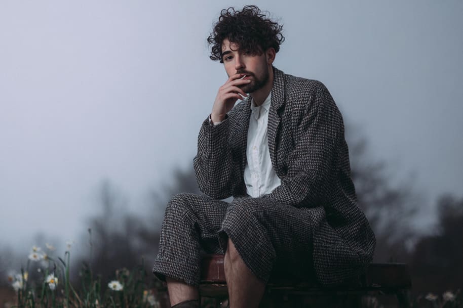 Stylish man with curly hair in a checkered suit, sitting outdoors on a foggy day, resting his hand on his chin with a contemplative expression, as one of new artists.