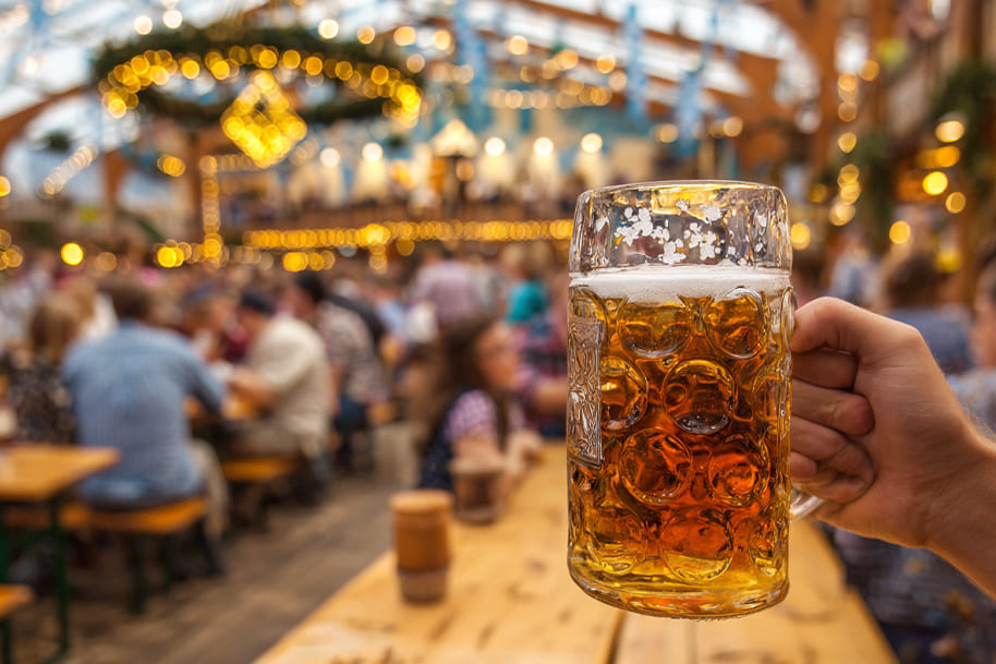 Close-up of a beer stein filled with golden Märzen beer held inside a festive Oktoberfest beer tent in Munich, with blurred crowd and lights in the background – featured on Eternal Slots Casino blog