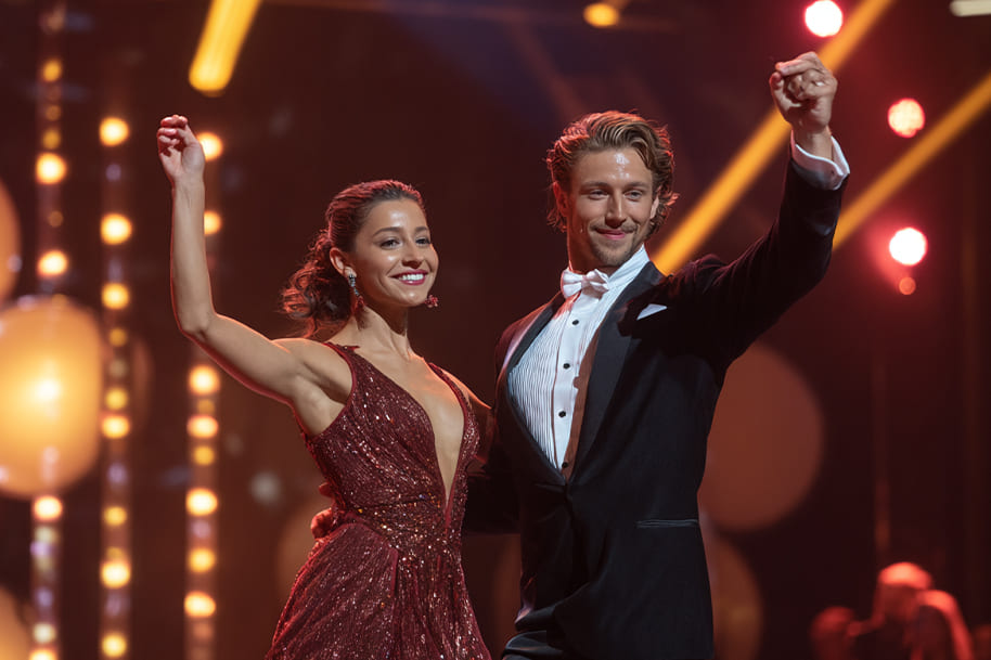 Professional dancers in a red dress and tuxedo taking a bow on stage under bright lights, celebrating a performance on Dancing with the Stars.