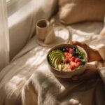A person sits comfortably by a window in soft morning light, holding a bowl filled with fresh avocado slices, strawberries, raspberries, blueberries, and grains. A warm cup of coffee rests nearby, creating a cozy and peaceful breakfast scene.