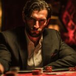 A man in a dark suit leans forward at a poker table under dim, golden lighting. His intense expression and steady posture reflect determination and focus during a high-stakes casino game.