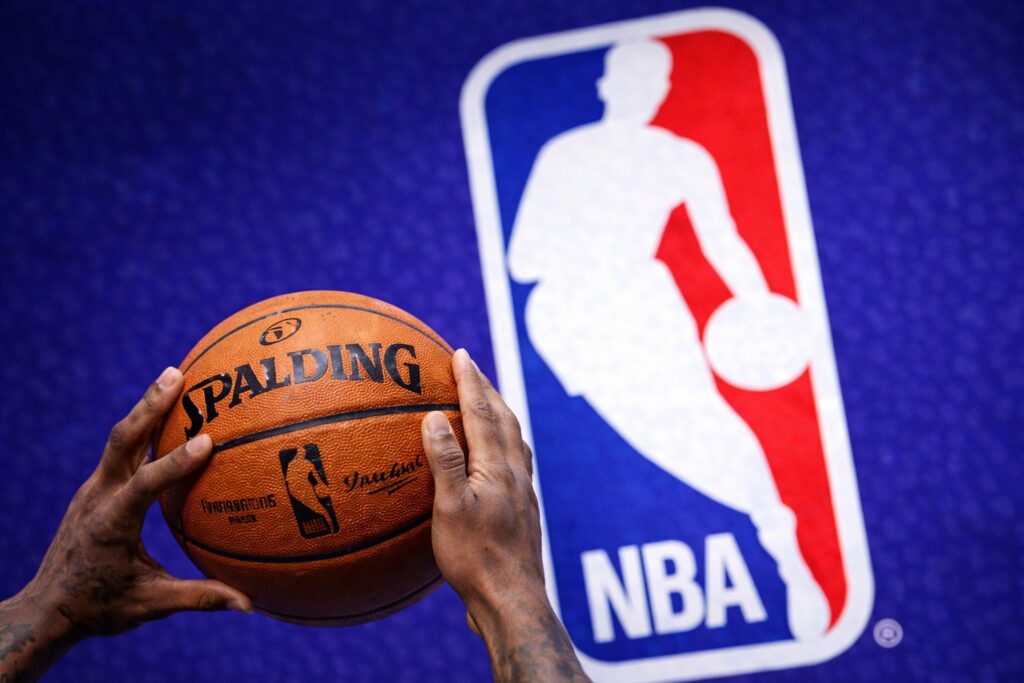 Close-up of hands holding an NBA basketball in front of a NBA logo background, symbolizing a league trade or game moment.
